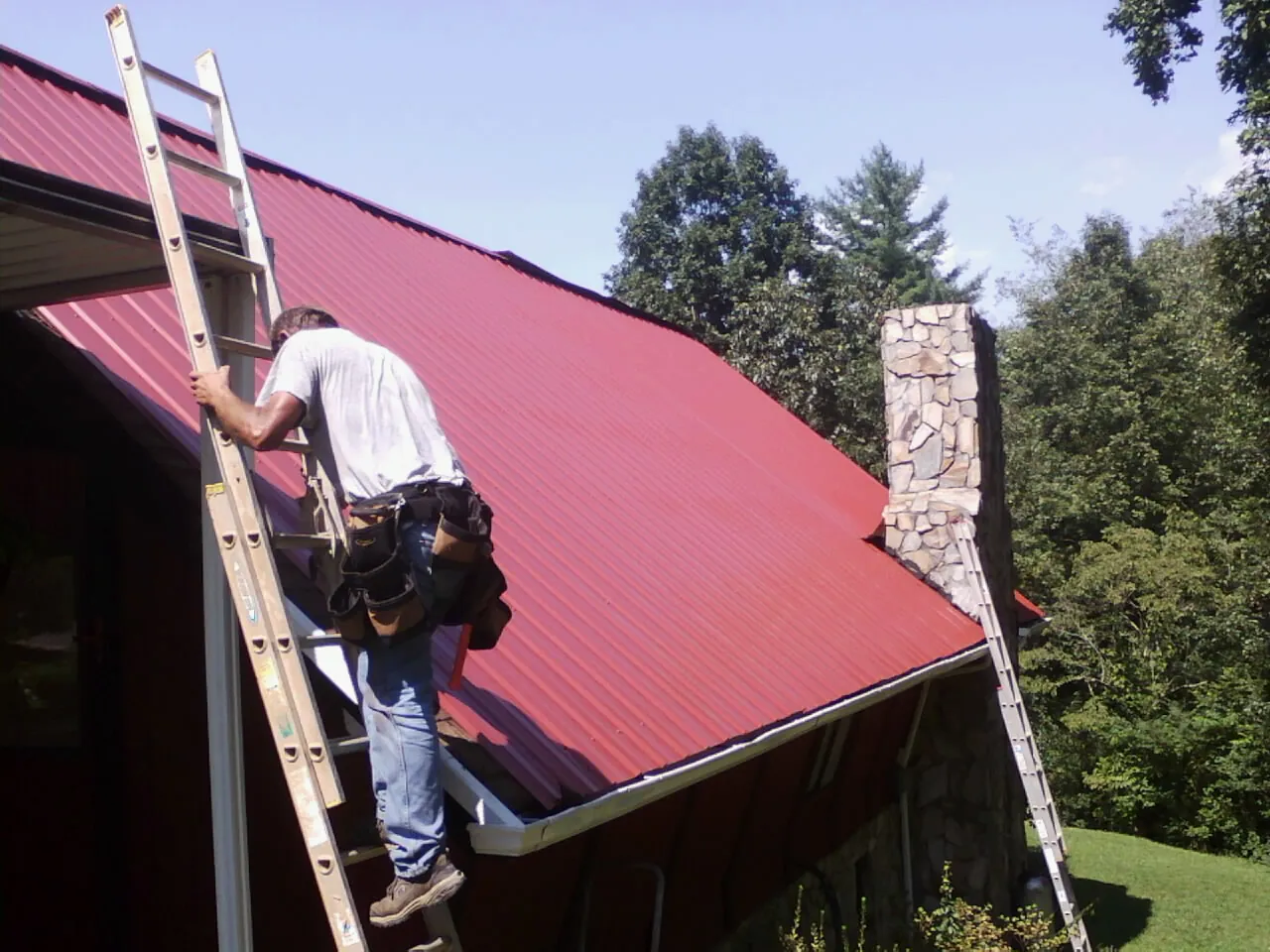 Roofer on a ladder beside a red metal roof and stone chimney — Soffit & Fascia Repair by Roofing Brothers in Escalante